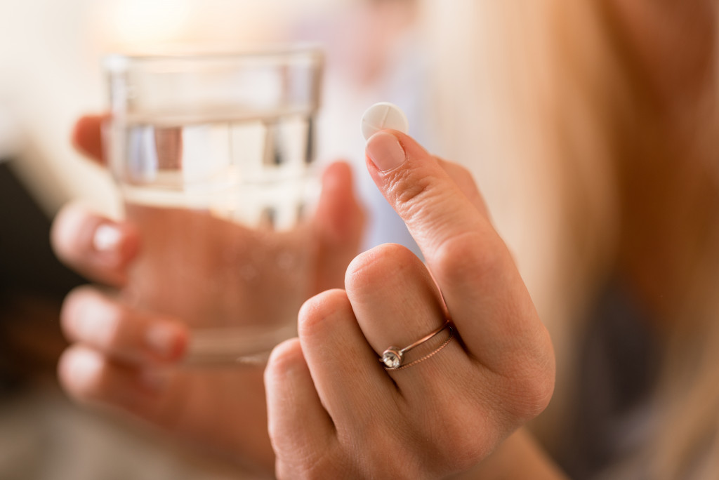 hand holding a pill in the foreground while holding a glass of water in the background